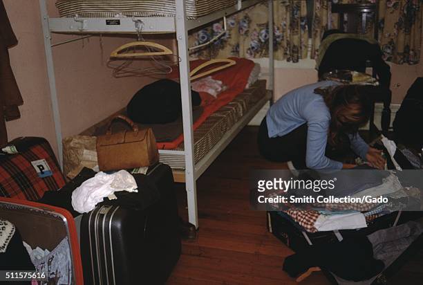 Photograph of a bedroom in a hostel, there are a large number of suitcases both open and closed throughout the room, a woman searches for something...