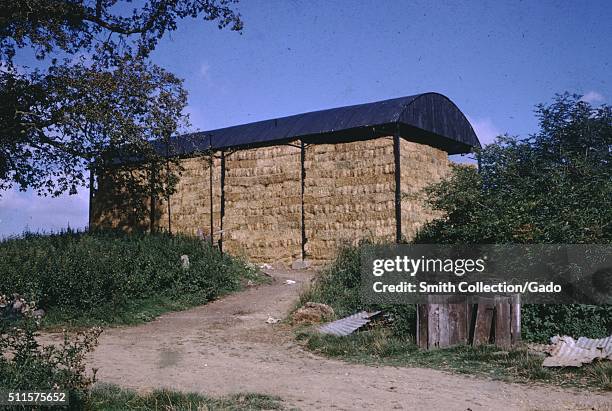 Photograph of a covered building with no sides used to store bales of hay, the bales are stacked several stories high, the building is situated on a...