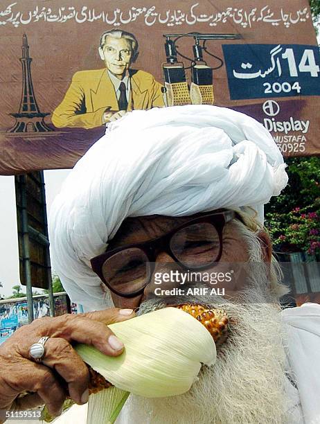 An elderly Pakistani man bites into a corn cob on a street where a billboard shows a protrait of country's first president Mohammad Ali Jinnah in...