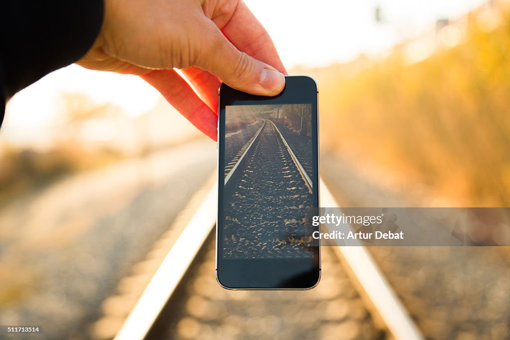 Man playing with perspective from personal point of view fitting smartphone screen on the railroad on sunset with nice vanishing point.