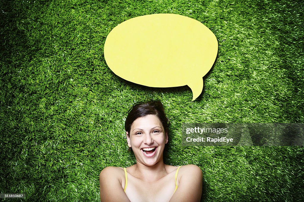 Pretty, laughing brunette lying on grass with blank speech bubble