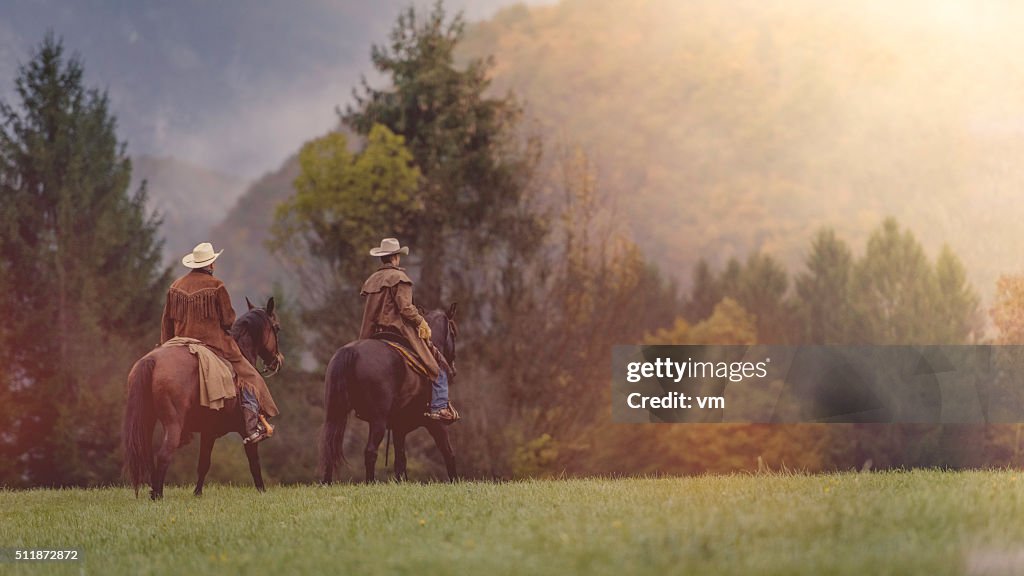 Two cowboys riding across a field in a forest