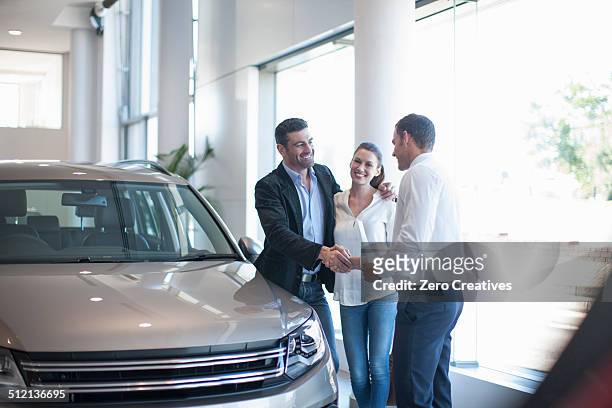 salesman shaking hands with mid adult couple in car dealership - dealer foto e immagini stock