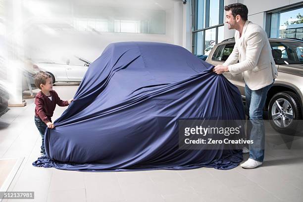 mid adult man uncovering new car with son in car dealership - new car stock pictures, royalty-free photos & images