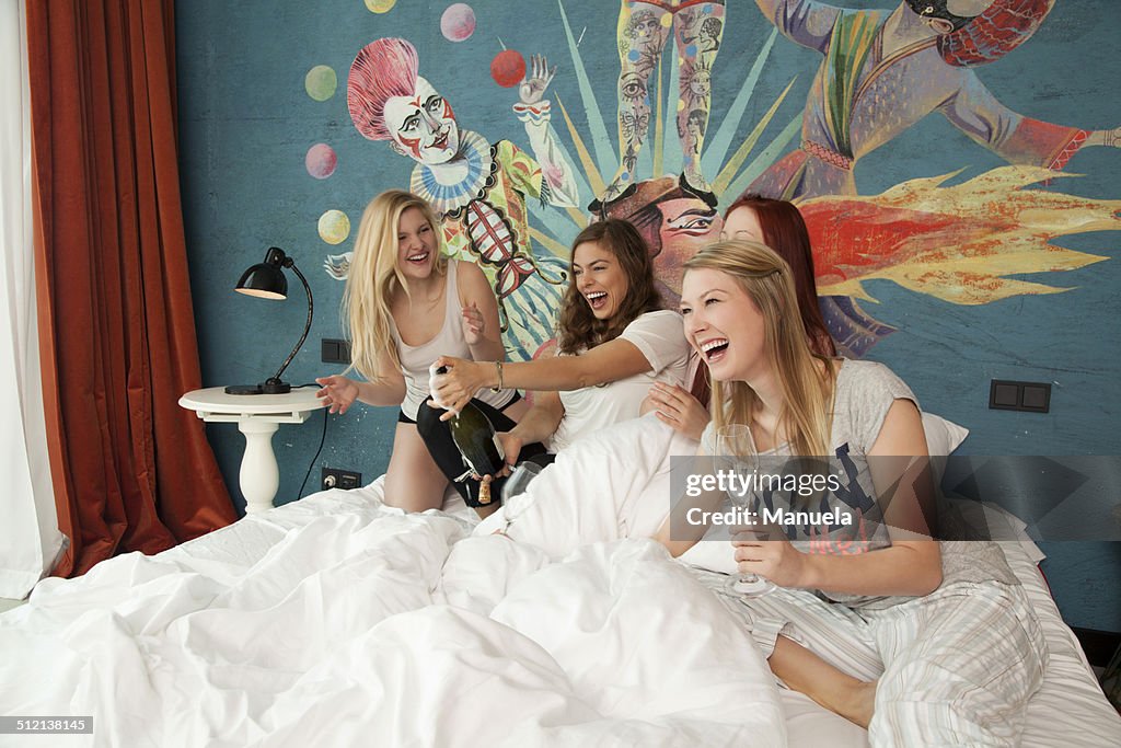 Four young women friends uncorking champagne on hotel bed