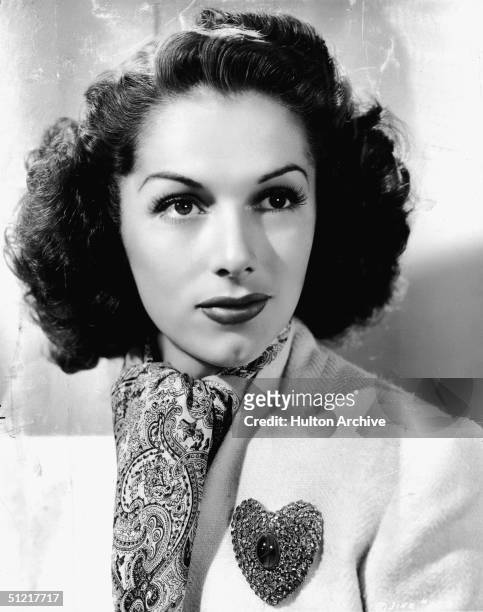 Portrait of Spanish-born American actress Jinx Falkenburg as she wears a white tweed suit, pink paisley scarf, and heart-shaped lapel pin set with a...