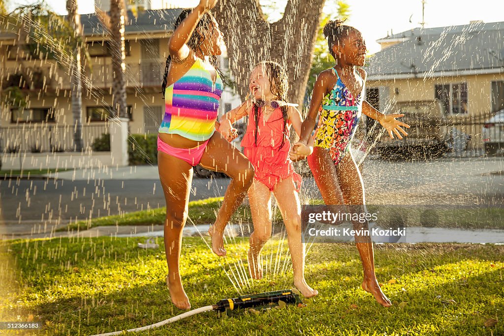 Three girls running and jumping in garden sprinkler