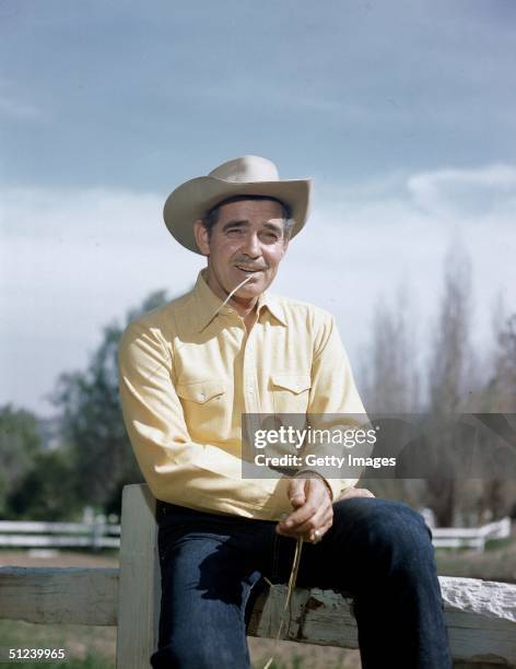 Circa 1945, American actor Clark Gable poses outdoors by a ranch fence, wearing a cowboy hat and chewing a stalk of wheat.