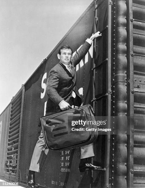 Circa 1965, American actor David Janssen hangs from the ladder of a freight train car while carrying a suitcase in still from the television show...