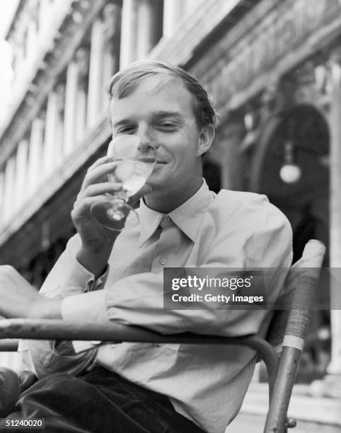 Circa 1955, American author Truman Capote smiles as he drinks a glass of ice water at an outdoor cafe in San Marco's Square Venice, Italy, 1950s.