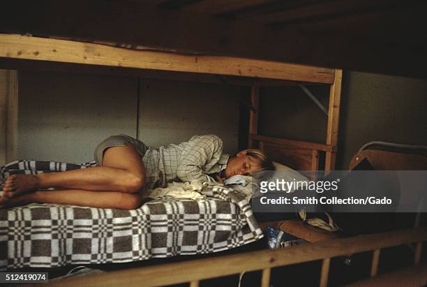 Young woman is shown sleeping on the lower portion of a bunk bed, she is fully clothed in a button down shirt and shorts, a chair containing some of...