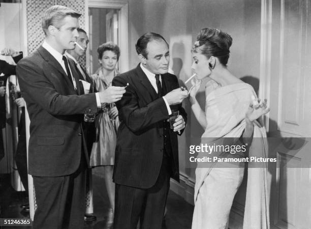 American actors George Peppard and Martin Balsam compete to light Belgian-born actor Audrey Hepburn's cigarette at a formal party in a still from...
