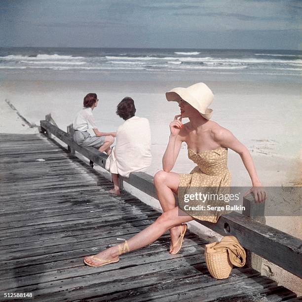 Model resting on the dock, wearing yellow petal-top bathing suit, corseted at the waist, straw hat, and sandals.