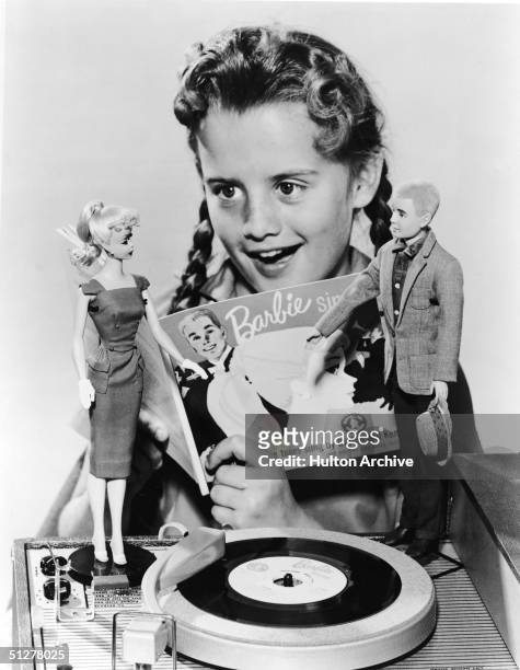 Girl in pigtails sings along with a 7" record called 'Barbie Sings' which plays on a portable phonograph player, 1961. Two dolls, Barbie and Ken,...