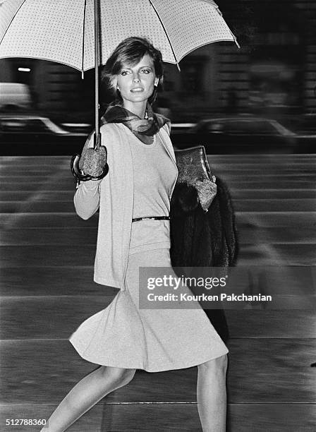 Model Cheryl Tiegs, walking under an umbrella by Mespo, holding a fur coat, and wearing a long angora and wool cardigan, matching sleeveless...
