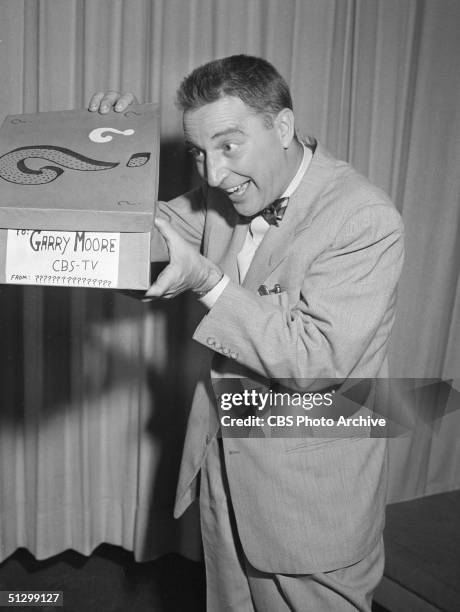 American television host Garry Moore peers excitedly into a box covered with question marks and labelled 'To Garry Moore, CBS-TV, from...