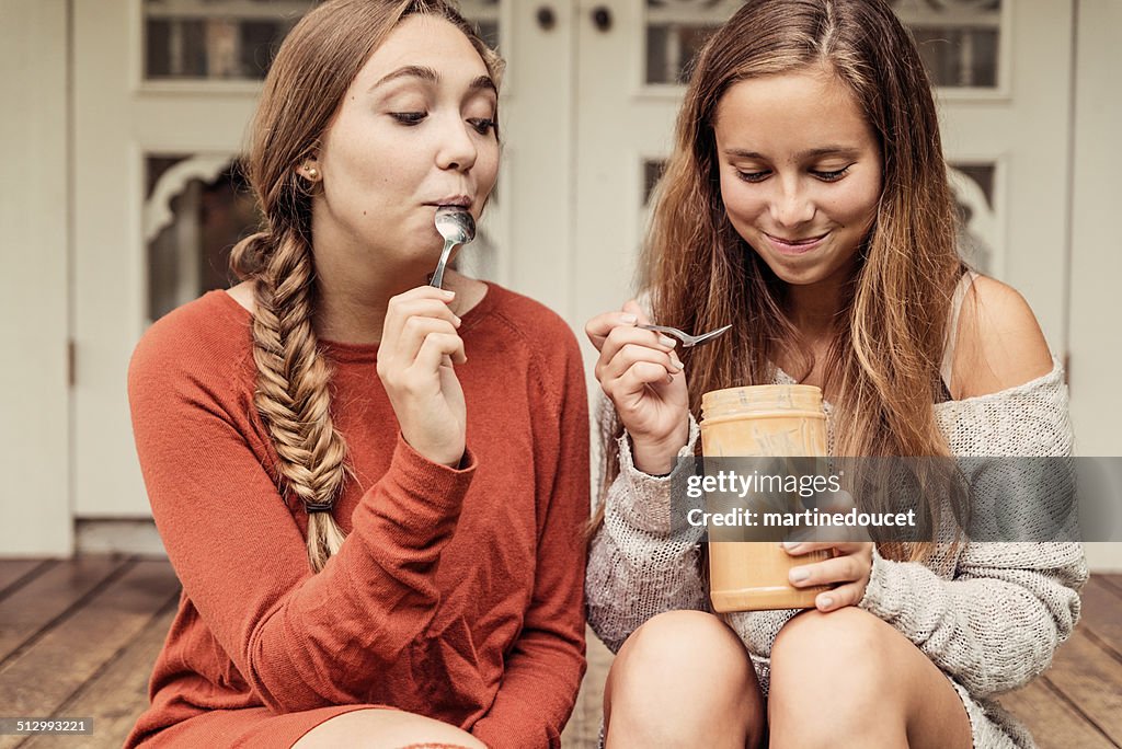 Teenage girlfriends eating peanut butter by the spoon on porch.