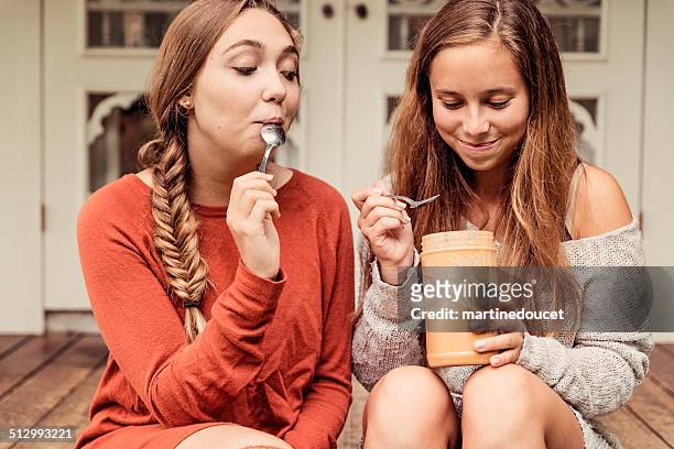 teenage girlfriends eating peanut butter by the spoon on porch. - licking lips stock pictures, royalty-free photos & images