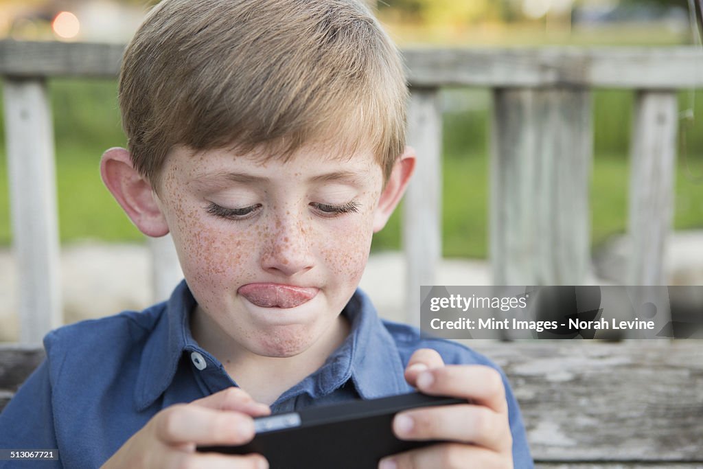 A young boy using a hand held electronic tablet or game and concentrating, sticking his tongue out.