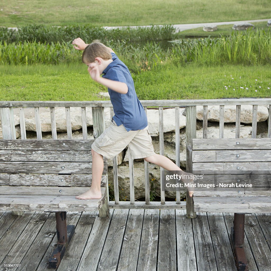 A young boy outdoors leaping from one bench to another on a jetty over water.