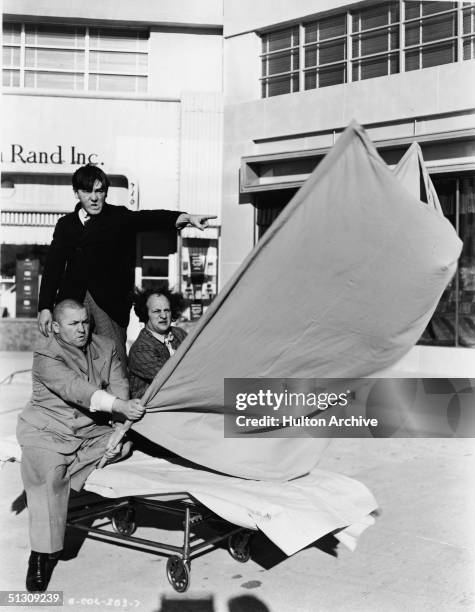 The Three Stooges ride a hospital gurney like a makeshift sailboat in a still from an unidentified film. L-R: American actors Curly Howard , Moe...