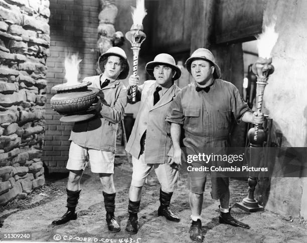 The Three Stooges, wearing safari outfits, hold torches as they explore a cave in a still from an unidentified film. L-R: American actors Larry Fine...