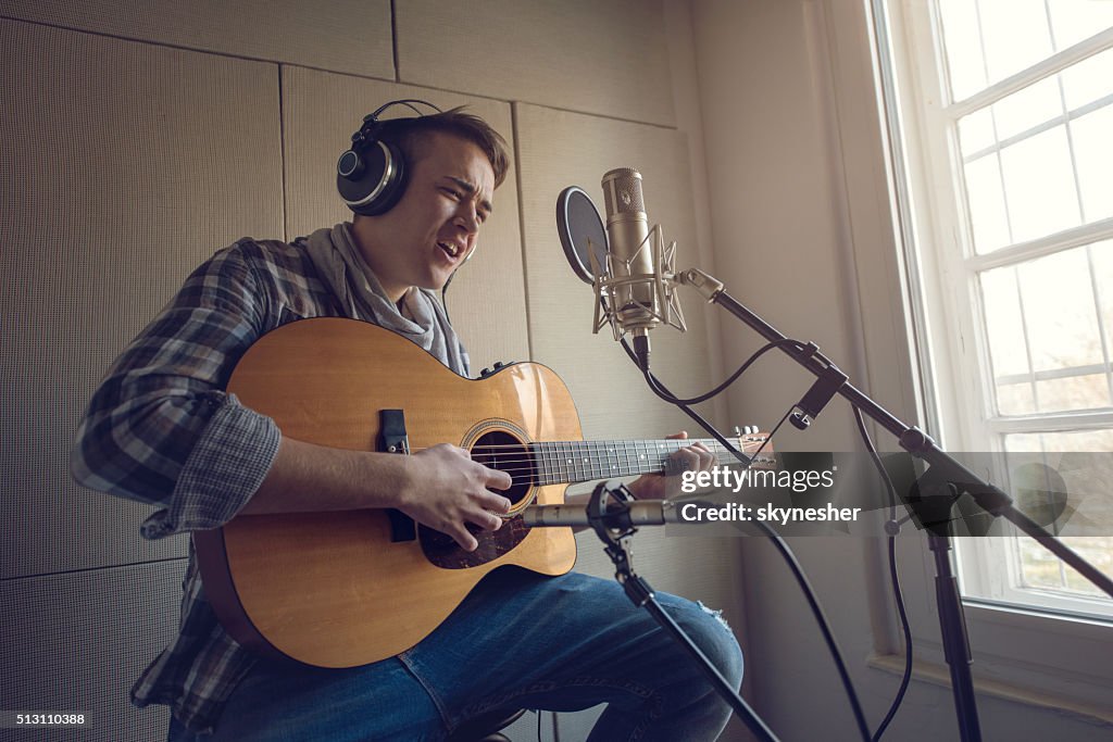 Young man playing acoustic guitar and singing in recording studio.