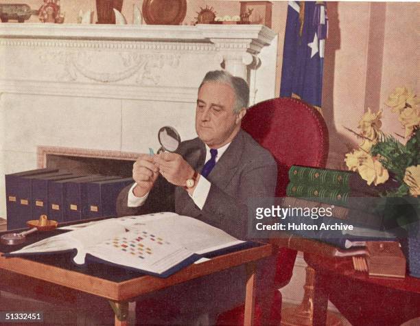 President Franklin Delano Roosevelt holds a stamp under a magnifying glass while seated at his desk with his stamp collection, Washington, DC, circa...