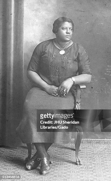 Full length seated portrait of young African American woman with neutral expression, wearing a darkly colored embroidered blouse, a dark skirt,...