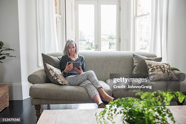 entspannende senior frau sitzt auf der couch mit digitaltablett - old woman sitting stock-fotos und bilder