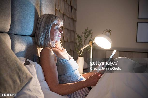 senior woman sitting in bed at night with tablet - bright bedroom stockfoto's en -beelden