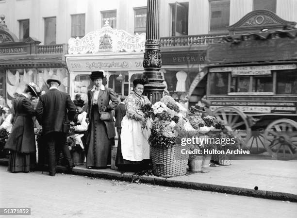 Woman sells flowers from a wicker basket beside a lamppost on Regent Street near Oxford Circus, London, 1911. Around her customers mill about other...