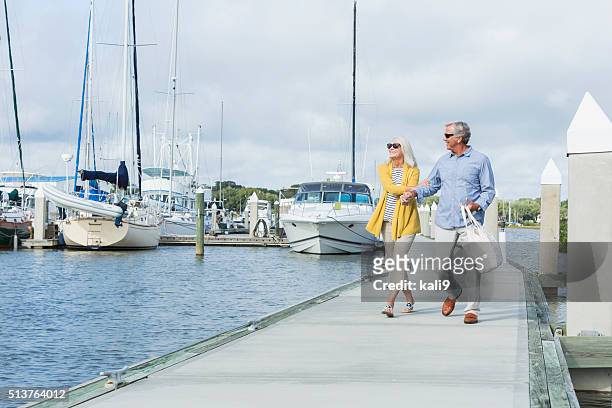 happy senior couple walking along harbor holding hands - couple boat stock pictures, royalty-free photos & images