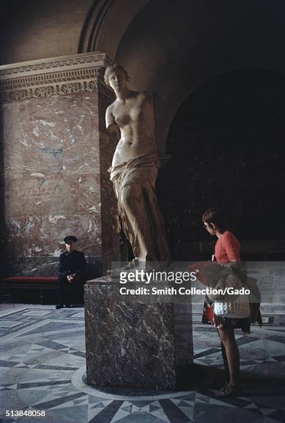 Photograph of the statue named Venus de Milo, the statue is believed to depict Aphrodite, a woman wearing a salmon colored top and carrying her coat...