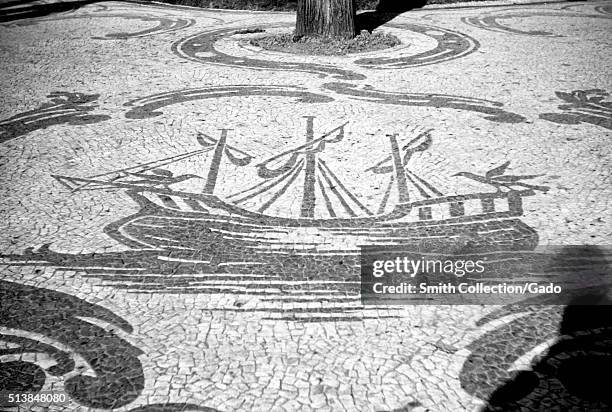 Photograph of a tile mosaic in an outdoor setting that wraps around a tree, the mosaic background is white tile while all the details are in a dark...