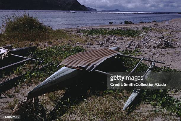 Outrigger canoe reinforced with a piece of corrugated steel, sitting in a patch of plants near a beach, American Samoa, 1961. .