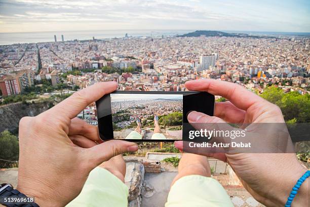 Man taking pictures from personal point of view with smartphone from the Turo de la Rovira hill with the Barcelona cityscape.