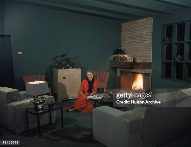 Portrait of American actress Janet Leigh as she sits on the floor of a green living room in a red skirt and jacket and reads a magazine at a coffee...