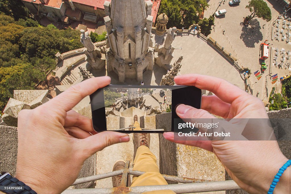 Traveller man taking pictures with smartphone from personal point of view of his legs on top of the Tibidabo church in Barcelona looking down.