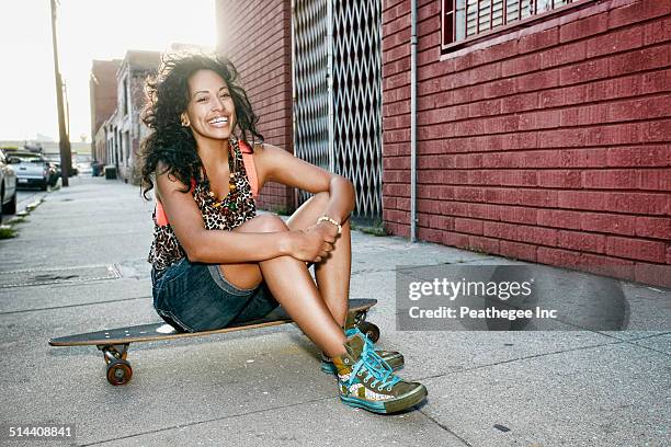 Hispanic woman sitting on skateboard on city street