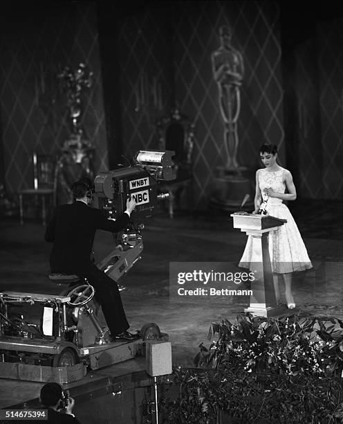 New York, NY: As a huge "Oscar" in the background looks down on the scene, Audrey Hepburn, with a TV camera filming the entire panorama, receives the...
