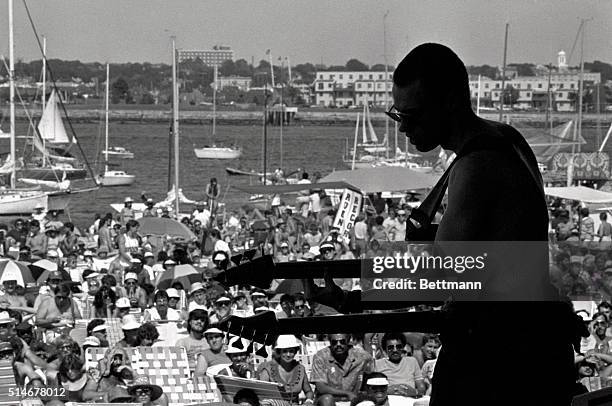 Musician Jonas Hellborg performs with the Mahavishnu Orchestra at the Newport Jazz Festival. A crowd relaxes in the sun along the harbor in Fort...