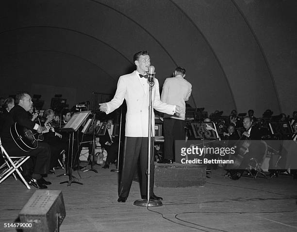 Frank Sinatra performs at the Hollywood Bowl. Here he stands full length crooning into a microphone, wearing a white jacket and black pants. SEE NOTE