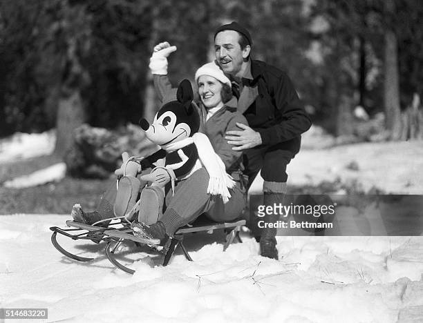 Mr. And Mrs. Walt Disney rest on a sled with a stuffed Mickey Mouse as they greet children to a snow party at the Lake Arrowhead resort area.