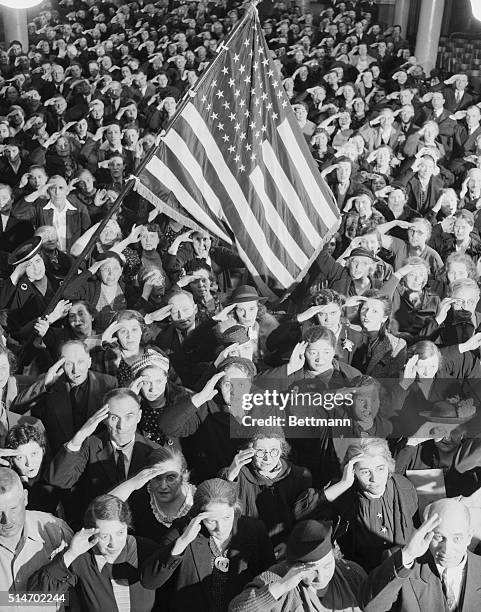 Night school students of all ages, many probably immigrants, stand in a large group saluting the American flag.