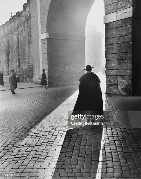 Rome, Italy: In pensive mood, a young priest walks slowly toward an arch in Rome's St. Peters Square, making a dramatic picture of late afternoon...