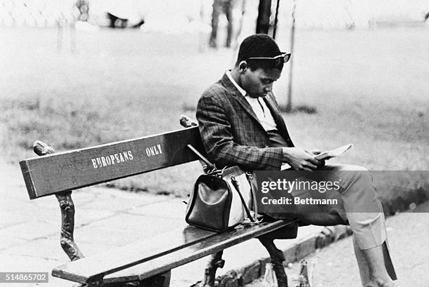 An African man reads on a bench marked "Europeans Only" in a South African park.