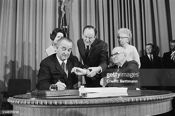 President Lyndon B. Johnson signs the Medicare bill as Vice President Humphrey and Harry S. Truman both check the time.