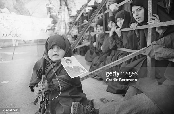 Tehran, Iran: A picture of Ayahtollah Ruhollah Khomeini pinned to her clothing, this little girl clutches a toy gun in front of the U.S. Embassy in...