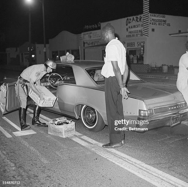 Los Angeles County Sheriffs search a car during the Watts Riots, finding a case of vodka, while the car's driver stands handcuffed nearby.